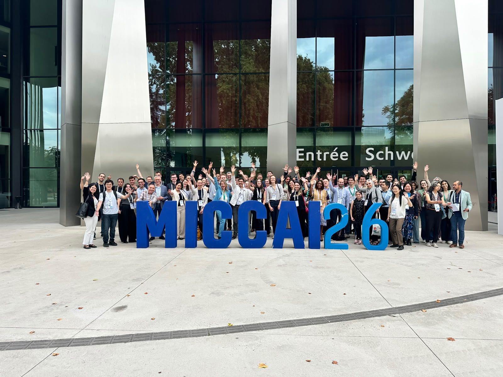 Group photo of MIART 2026 participants waving behind large blue MICCAI26 letters outside the Strasbourg convention centre